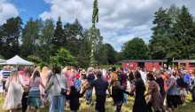 Traditionell ringdans runt en midsommarstång framför ett tåg på en museijärnväg.