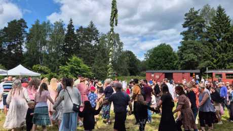 Traditionell ringdans runt en midsommarstång framför ett tåg på en museijärnväg.